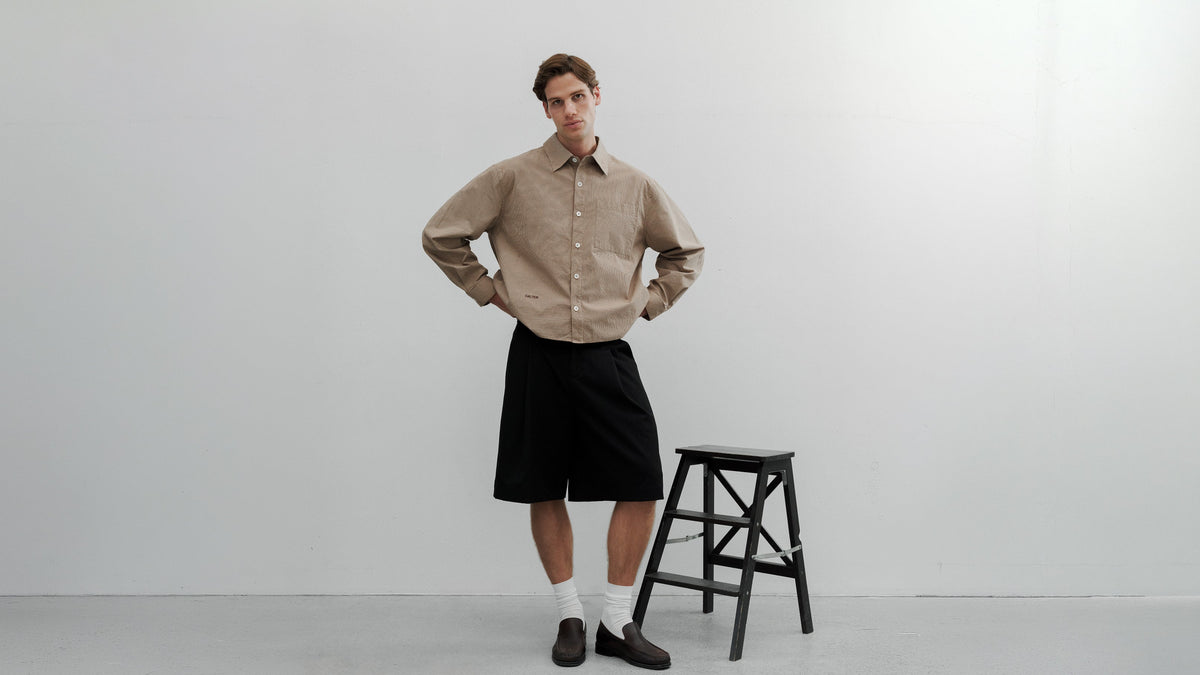 Person wearing a beige shirt and black shorts standing next to a black stool on a white background