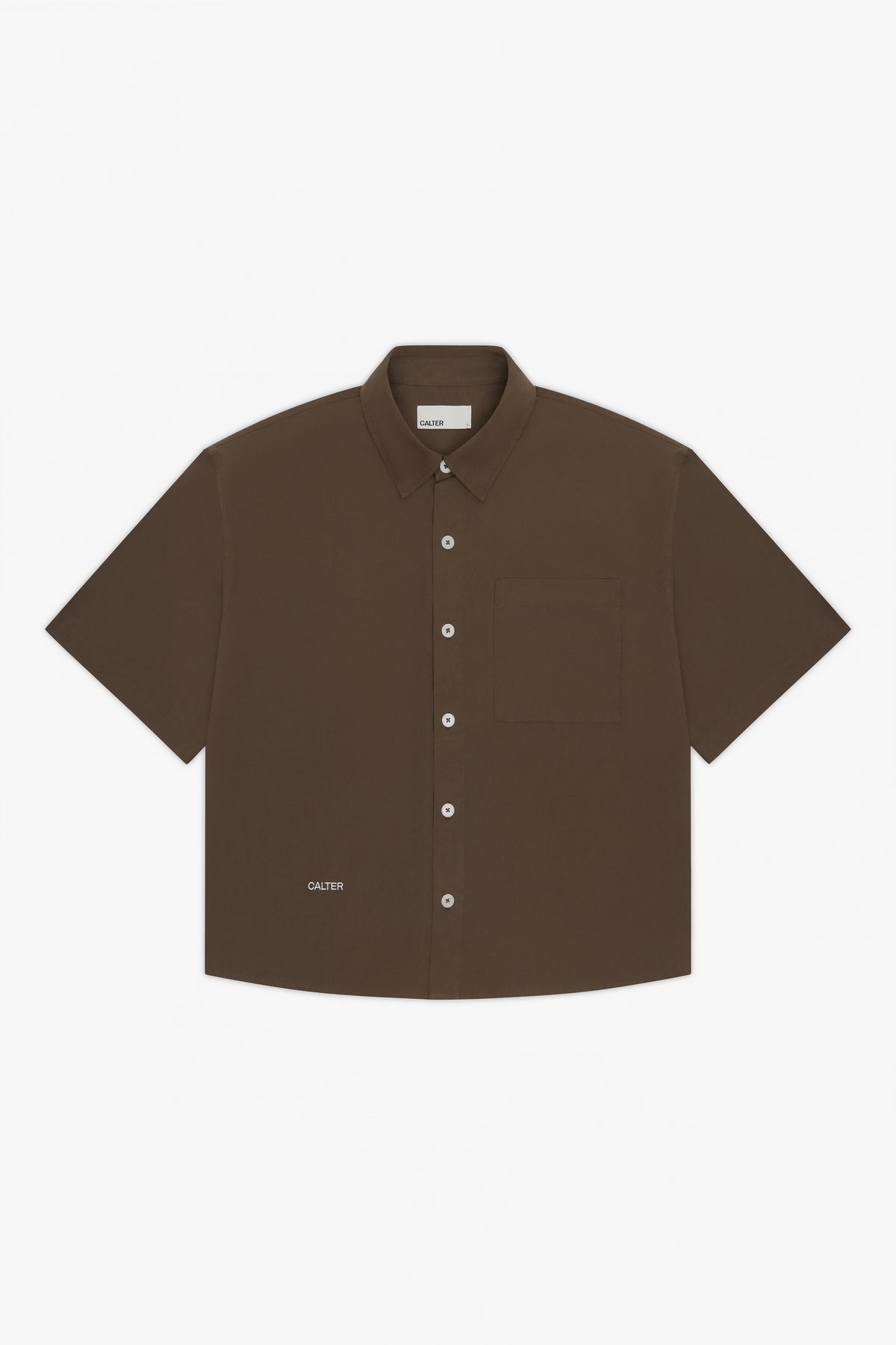 The Calter Volume Shirt is a short-sleeved, dark brown cotton poplin shirt with a boxy fit, single chest pocket, white buttons, and a small CALTER logo near the bottom hem, shown laid flat on a white background.