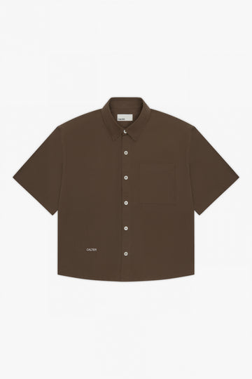 The Calter Volume Shirt is a short-sleeved, dark brown cotton poplin shirt with a boxy fit, single chest pocket, white buttons, and a small CALTER logo near the bottom hem, shown laid flat on a white background.
