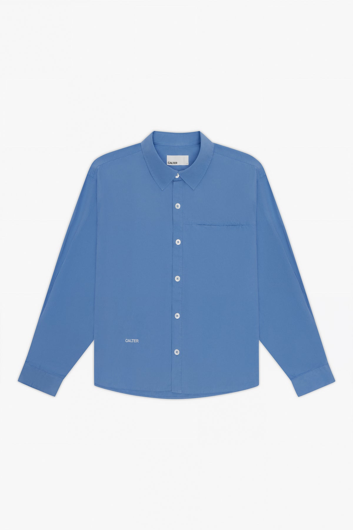 The Calter Pleated Shirt is a long-sleeved, boxy button-up in solid blue cotton poplin with white buttons and a left chest pocket, shown on a plain white background.