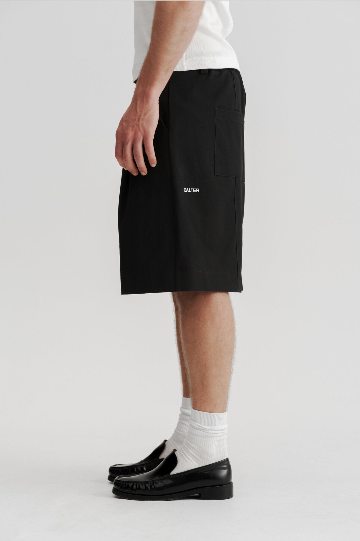 Side view of a model wearing Black Pleated Shorts, showing the below-knee length and rear CALTER embroidery.