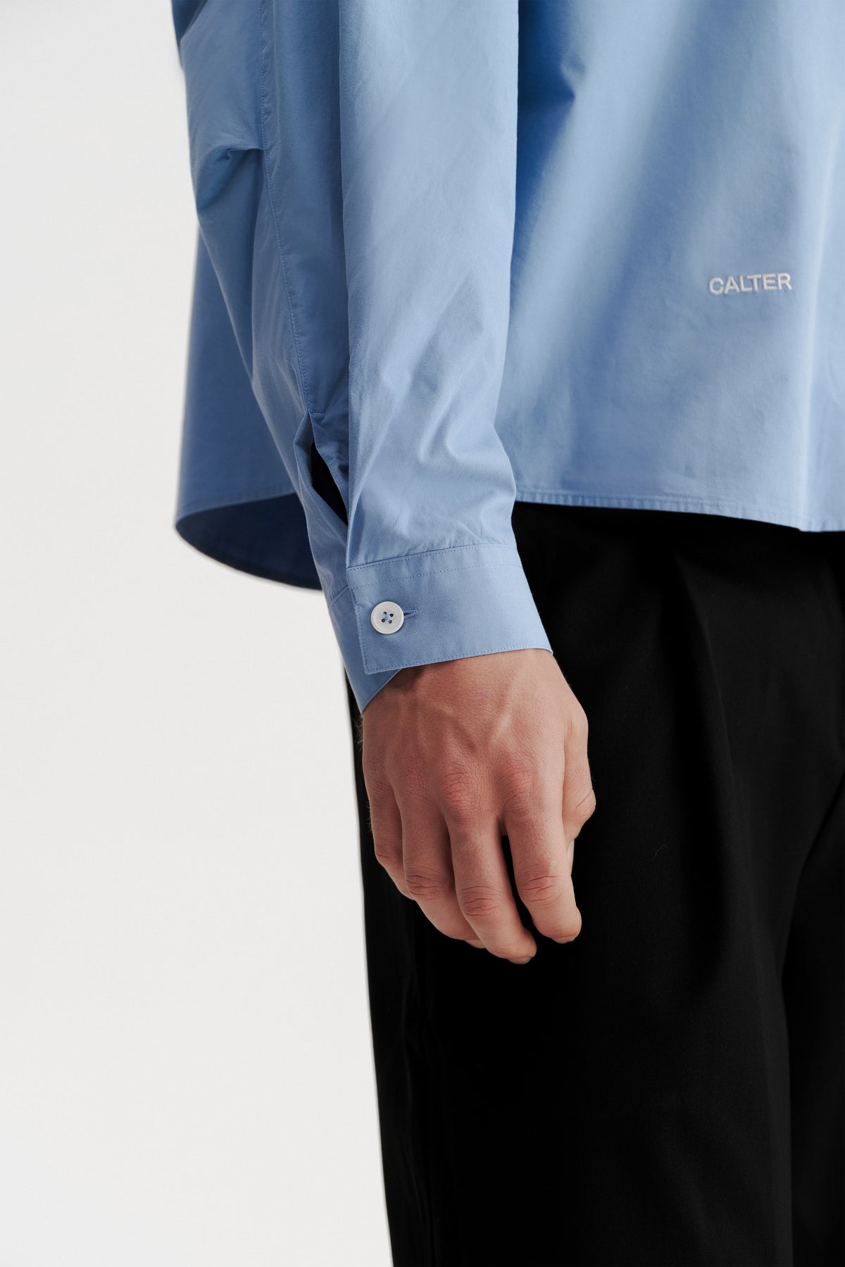 Close-up of the buttoned cuff, wrist pleat and front-hem embroidery on the Technical Blue Pleated Shirt.