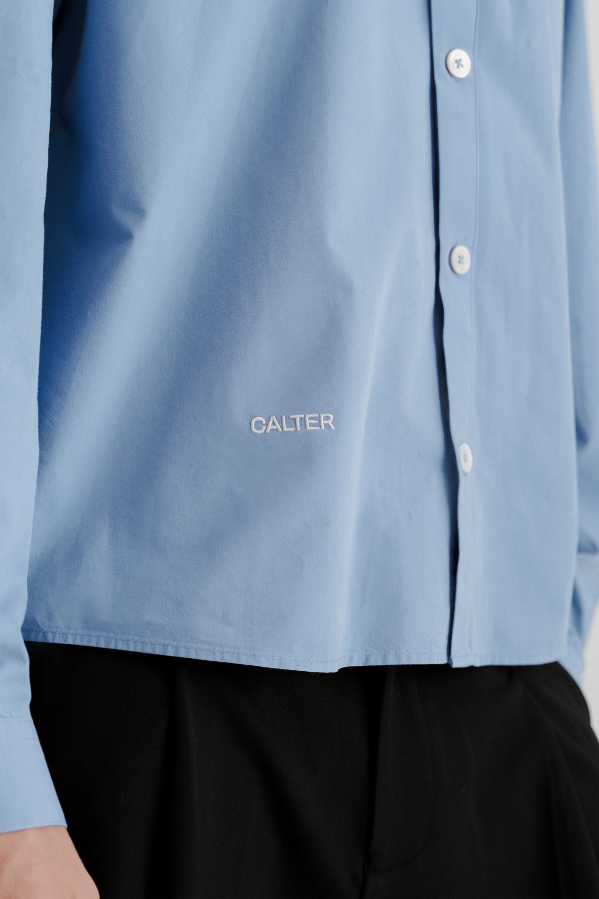 Close-up of the white CALTER embroidery near the front hem of the Technical Blue Pleated Shirt.