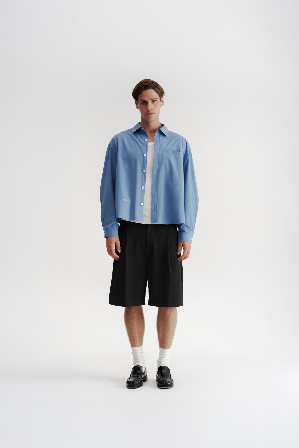 Full-length front view of a model wearing the Technical Blue Pleated Shirt open over a white tank top.