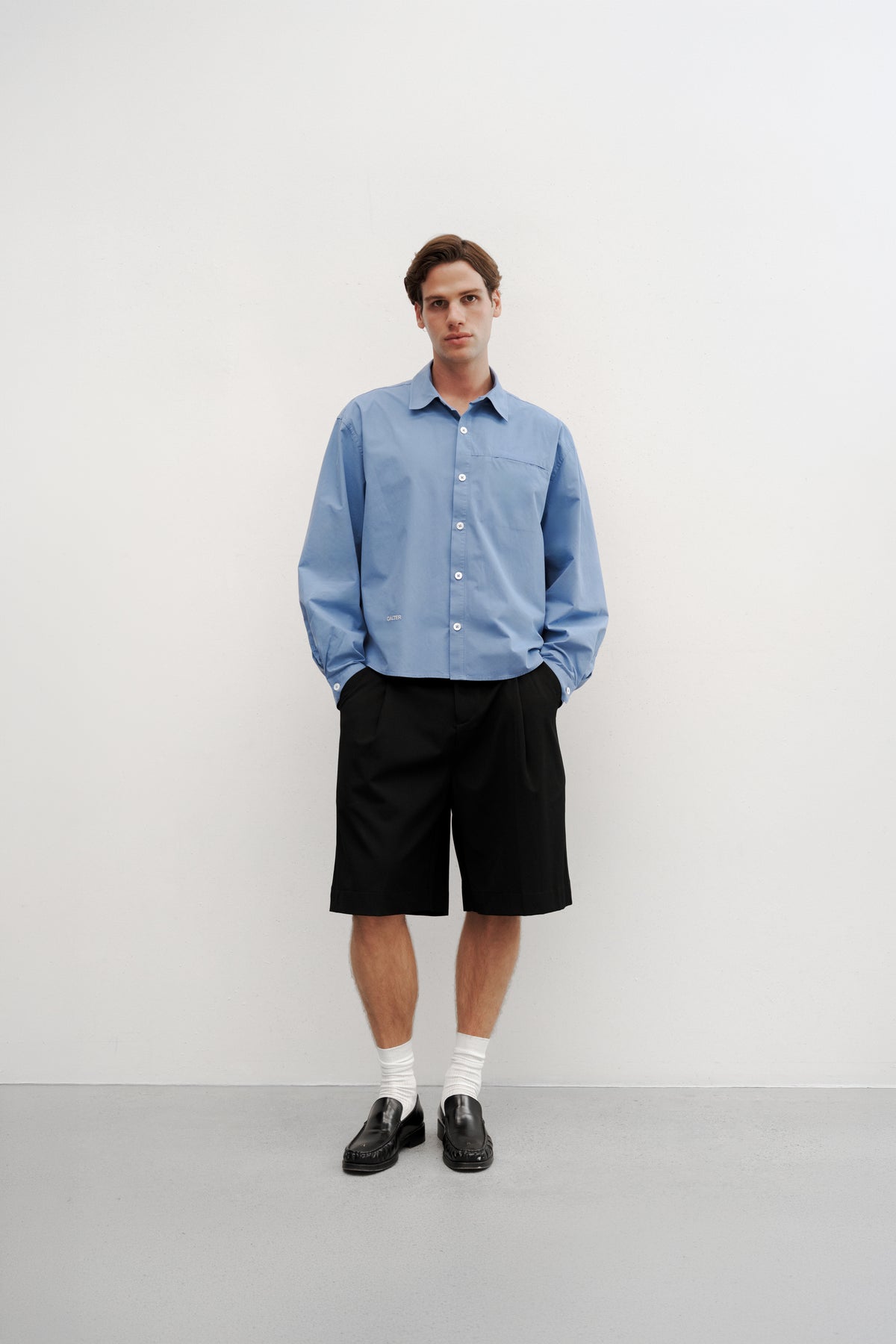 Full-length front view of a model wearing the Technical Blue Pleated Shirt buttoned with Black Pleated Shorts.