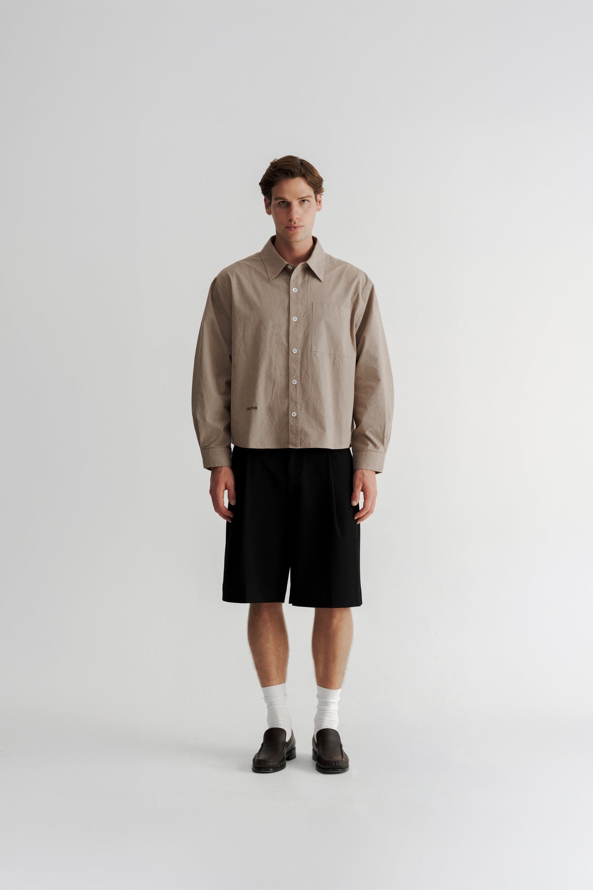 Full-length front view of a model wearing the Sand Microcheck Studio Shirt with Black Pleated Shorts.