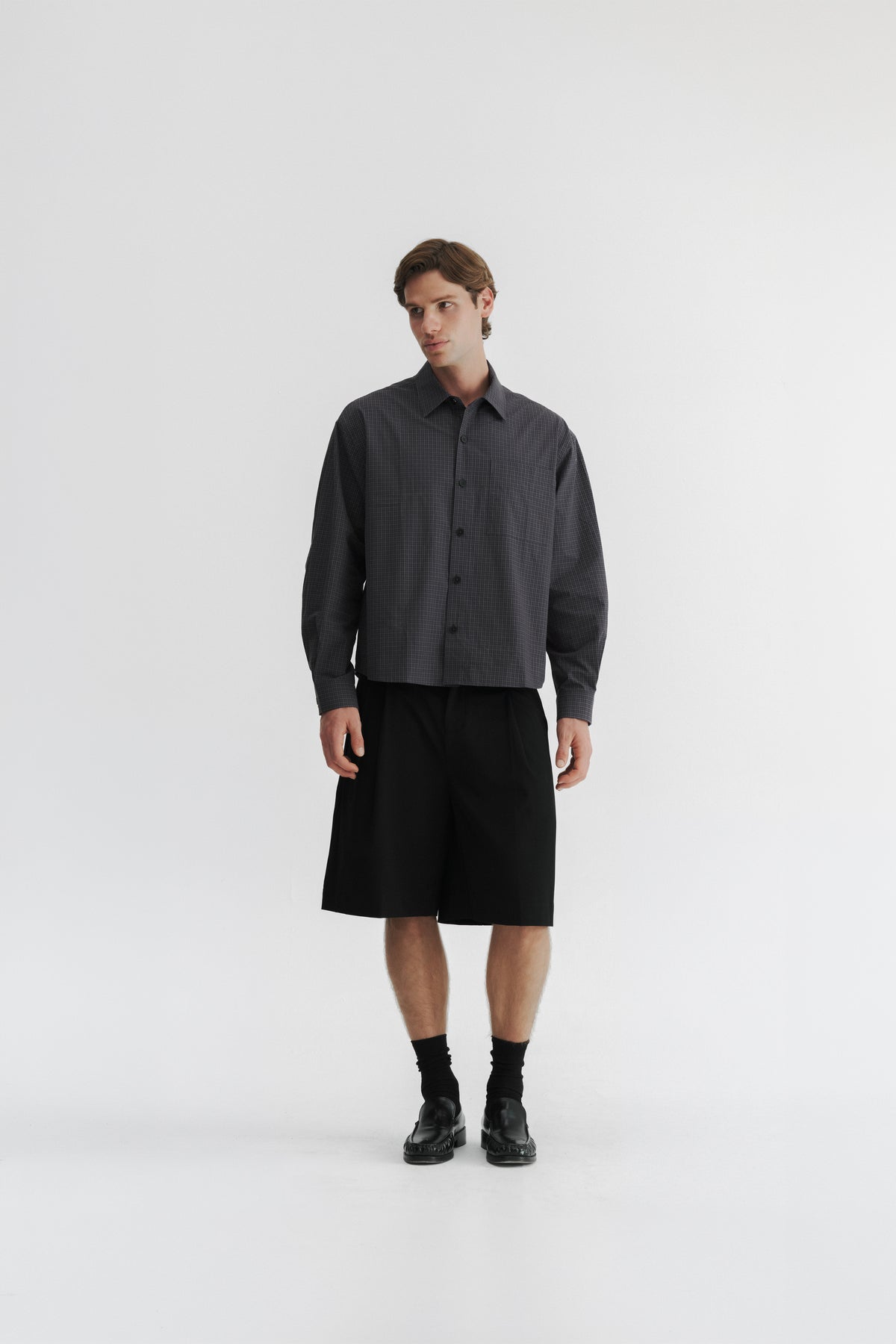 Three-quarter front view of a model wearing the Slate Microcheck Studio Shirt with Black Pleated Shorts.