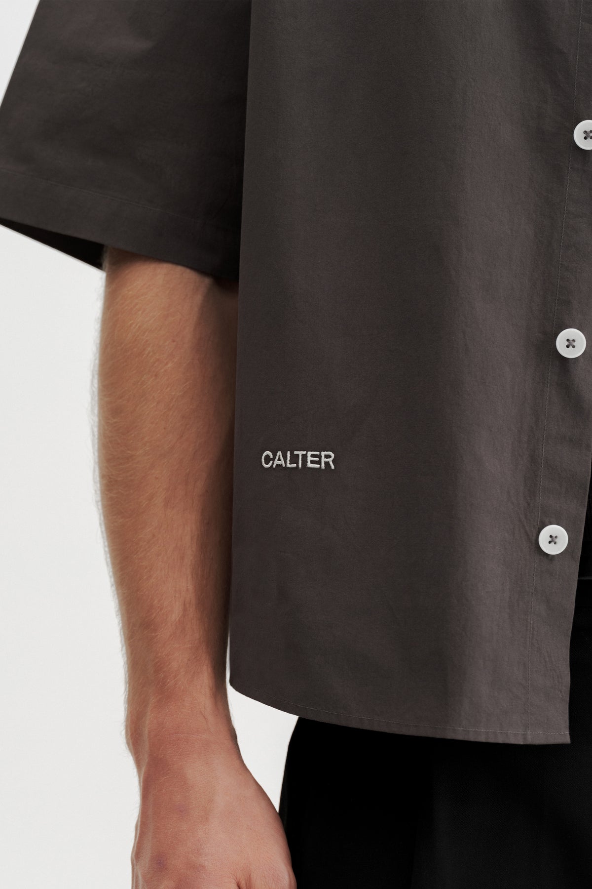 Close-up of the buttons and white CALTER embroidery near the front hem of the Charcoal Volume Shirt.