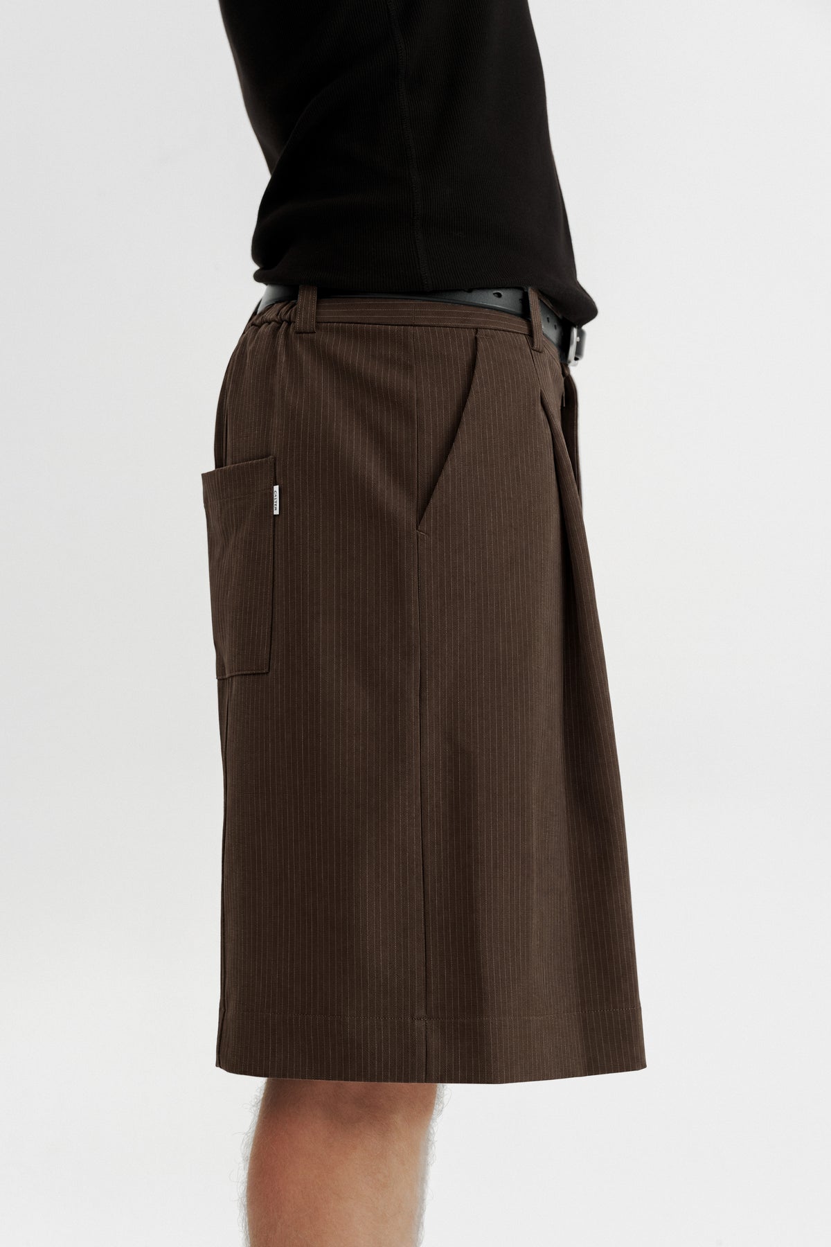 Side view of a model wearing Brown Stripe Pleated Shorts, showing the fine pinstripe and rear patch pocket.